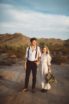 a man standing next to a woman in a white dress on a dirt road holding a bottle