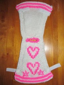 a white knitted mitt with pink hearts on the front and bottom, sitting on a wooden floor