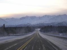 an empty road with snow on the ground and mountains in the background