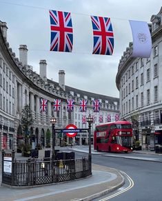 a red double decker bus driving down a street next to tall buildings with flags hanging from it's sides