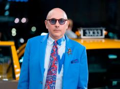 a bald man in a blue suit and tie standing next to a yellow taxi cab