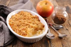 an apple crumbler in a white dish next to two spoons on a wooden table