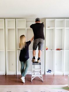 a man and woman are standing on a ladder in front of bookshelves while they paint the walls