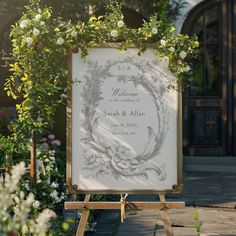 a wedding sign with greenery around it and flowers on the frame, in front of a building