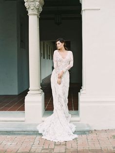 a woman standing in front of a white building wearing a long sleeved wedding dress