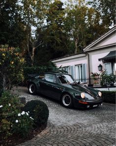 a black car parked in front of a house on a cobblestone driveway next to bushes and trees