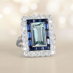 This item has just been reserved by another customer. Please contact us to be notified if it becomes available.  This stunning 5ct ring showcases a rectangular aquamarine gem that has been carefully cut in an emerald shape. The 5ct ring aquamarine is securely placed in a protective setting and is encircled by two layers of sparkling sapphires and diamonds. The sides of the 5ct ring are embellished with a beautiful design of diamonds in the shape of a lemur-de-lis. Made with skillful craftsmanshi Art Deco Rectangular Emerald Ring For Anniversary, Luxury Blue Rectangular Sapphire Ring, Rectangular Topaz And Diamond Ring For Anniversary, Rectangular Diamond Topaz Ring For Anniversary, Rectangular Blue Topaz Ring With Diamond Accents, Blue Rectangular Diamond Ring, Elegant Multi-stone Rectangular Rings, Luxury Rectangular Diamond Ring With Accent Stones, Gia Certified Rectangular Sapphire Ring For Anniversary