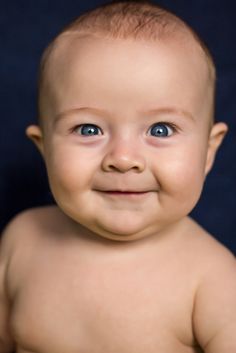 a smiling baby with blue eyes and no shirt on, looking at the camera while sitting in front of a black backdrop