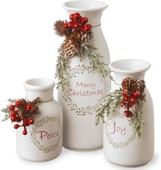 three white vases decorated with holly and pine cones, one has a merry christmas message on it