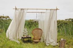 an old chair sitting in the middle of a lush green field next to a white curtain