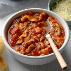 a white bowl filled with pasta and meat soup next to bread on a table top