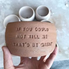 a hand holding up a clay container with words on it and four white cups in the background