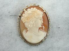 Versatile and lovely, this pendant is crafted of fine 18k gold. The frame work is simple and scalloped, with lovely, detailed motifs surrounding the solid, simple bezel frame. The cameo itself is perfectly carved, a lady in profile wearing a braided crown, earrings and necklace. The color of the shell is a rich, warm tan, and the carved cameo portion is creamy with hints of coral. Cameo is the art of carving a layered material, creating a portrait or landscape with the difference in the colors. Elegant Cameo Jewelry For Wedding, Classic Cameo Brooches For Wedding, Engraved Yellow Gold Pendant Brooch, Elegant Yellow Gold Baroque Brooches, Classic Gold Brooch With Intaglio Detail, Elegant Medallion Brooch For Collectors, Classic Gold Brooches With Intaglio, Classic Gold Intaglio Brooch, Elegant Cameo Jewelry