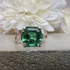 an emerald colored ring with two white diamonds on the side and a flower in the background