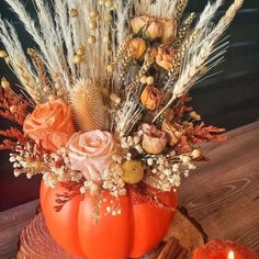 an arrangement of flowers and plants in a pumpkin shaped vase on a wooden table next to two candles