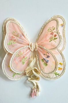 a pink butterfly shaped wall hanging with tassels on it's sides and an embroidered ribbon around the wings