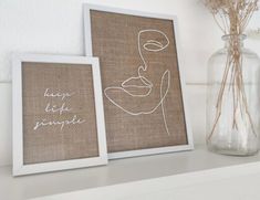 two framed pictures sitting on top of a white shelf next to a vase with dried flowers