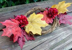 a wreath with red, yellow and green leaves is sitting on a wooden bench next to flowers