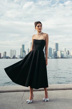 a woman in a black dress is standing near the water and looking at the camera