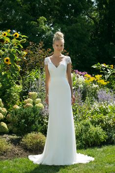 a woman in a white wedding dress standing on the grass with flowers behind her and looking at the camera