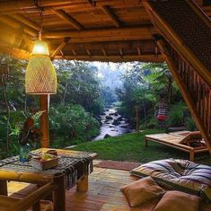 an outdoor living area with wooden furniture and plants on the side of the house, overlooking a stream