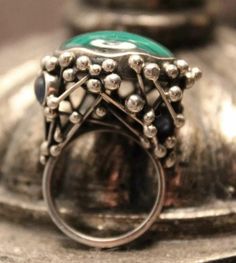 Add a unique touch to your jewelry collection with this stunning handmade abstract ring. The ring features a beautiful cabochon-shaped malachite gemstone, set in sterling silver. The green-colored ring is a perfect accessory for any occasion. The ring is crafted in the United States and has a ring size of 8. The secondary stone, sapphire, adds to the uniqueness of the ring. The malachite cabochon is the main attraction, but the sapphire stone adds an extra sparkle to the piece. The ring is perfect for those who appreciate fine jewelry and want to add a unique touch to their collection. Unique Green Polished Turquoise Ring, Unique Green Turquoise Ring With Polished Finish, Green Emerald Cabochon Rings, Unique Oval Cabochon Emerald Ring, Green Gemstone Cabochons Fine Jewelry, Green Cabochon Ring Jewelry, Elegant Green Chrysocolla Jewelry, Handmade Green Oval Cabochon Ring, Silver Emerald Cabochon Ring