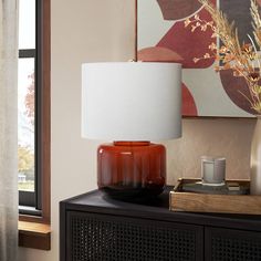 a table lamp sitting on top of a wooden cabinet next to a painting and window