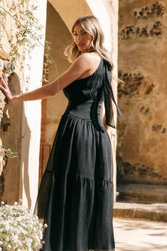 a woman in a black dress is standing by a wall with flowers and greenery