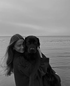 a woman holding a black dog on the beach near the ocean with her eyes closed