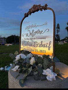 a welcome sign sitting on top of a cement slab with flowers in the foreground