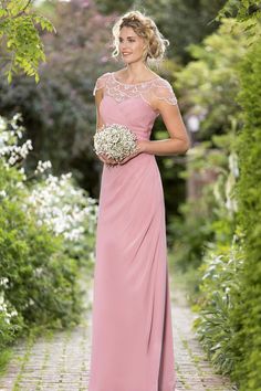 a woman in a long pink dress standing on a brick path with flowers and greenery behind her