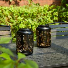 two black candles sitting on top of a wooden table next to green plants and bushes