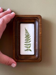 a hand is holding a small wooden frame with a green fern print on the inside