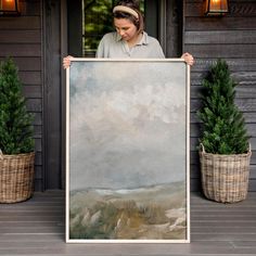 a woman holding up a large painting in front of a house with potted plants