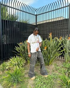 a man standing in front of a black fence with flowers and plants around him on the ground