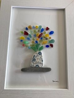 a white frame holding a glass vase with multicolored beads in it and a rock underneath