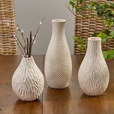 three white vases sitting on top of a wooden table next to plants and wicker baskets