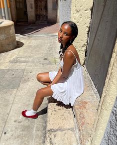 a young woman sitting on the side of a building wearing red shoes and a white dress