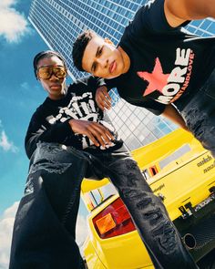 two young men standing next to each other in front of a yellow truck and tall buildings