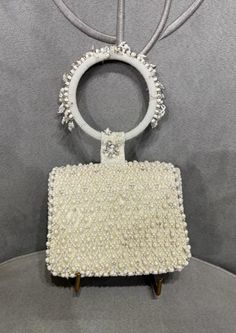 The Joel design pays homage to heritage jaali work, capturing its mysterious play of light and shadow through intricate pearl and beadwork. Natural pearl drops enhance its graceful demeanor. Our mini trapezium bag is a compact and edgy silhouette. This flap over is designed with soft edges. The embellished attached round handle makes it a statement hand held bag. This collectible accessory fits in your valuables like makeup, keys, currency and cards. Elegant White Rectangular Jewelry, Traditional White Jewelry For Evening, Elegant Embellished Jewelry For Events, Formal Pearl Embellished Jewelry, Formal Embellished Pearl Jewelry, Traditional White Evening Jewelry, Luxury White Beaded Jewelry, Party Jewelry With Pearl Embroidery, Elegant Beige Wedding Jewelry