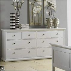 a white dresser with two paintings on the wall and flowers in vases next to it