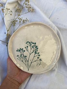 a hand holding a white plate with flowers on it and some fabric in the background