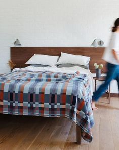 a woman walking across a wooden floor next to a bed in a white walled room