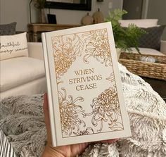 a person holding up a book in front of a white couch with pillows on it