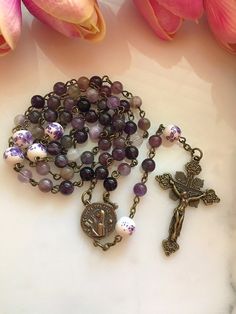 "Handmade bronze St. Teresa of Avila rosary made with 6mm purple amethyst gemstone beads and 8mm purple & white floral ceramic beads. The vintage inspired bronze Saint Teresa center measures 13/16\". The crucifix measures approximately 1 3/4\"." Lavender Spiritual Jewelry With 8mm Beads, Spiritual Lavender Jewelry With 8mm Beads, Lavender Spiritual Jewelry With Beads, Purple Rosary With 8mm Beads, Purple Rosary With 8mm Round Beads, Handmade Adjustable Purple Rosary, Handmade Spiritual Purple Rosary Bracelet, Purple Beaded Rosary As Gift, Beautiful Rosaries