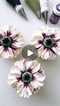 three white and purple flowers sitting on top of a table next to some paint bottles