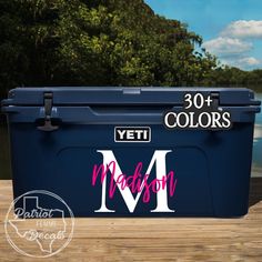 a blue cooler with the letter m on it sitting on a wooden deck next to water