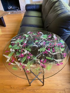 a glass table with flowers on it in front of a black couch and wooden floor