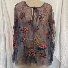 Sheer Mesh Blouse With Intricate Floral Embroidery. The Blouse Has Long Sleeves And A Round Neckline That Ties At The Front. The Fabric Is Lightweight And Transparent, With A Colorful Floral Pattern That Includes Red, Pink, Yellow, And Blue Flowers. The Embroidery Is Detailed And Appears To Be Done With Threads Of Various Colors, Giving The Blouse A Vibrant And Textured Look. The Blouse Also Features Ruffled Cuffs And A Loose, Flowy Silhouette. Bohemian Embroidered Silk Tops, Bohemian Silk Embroidered Tops, Purple Bohemian Top With Floral Embroidery, Bohemian Purple Top With Floral Embroidery, Festive Purple Tops With Resham Embroidery, Spring Purple Embroidered Blouse, Purple Floral Embroidered Top For Spring, Spring Embroidered Purple Blouse, Bohemian Silk Blouse With Floral Embroidery