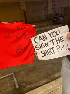 a woman holding up a sign that says can you sign the shirt?